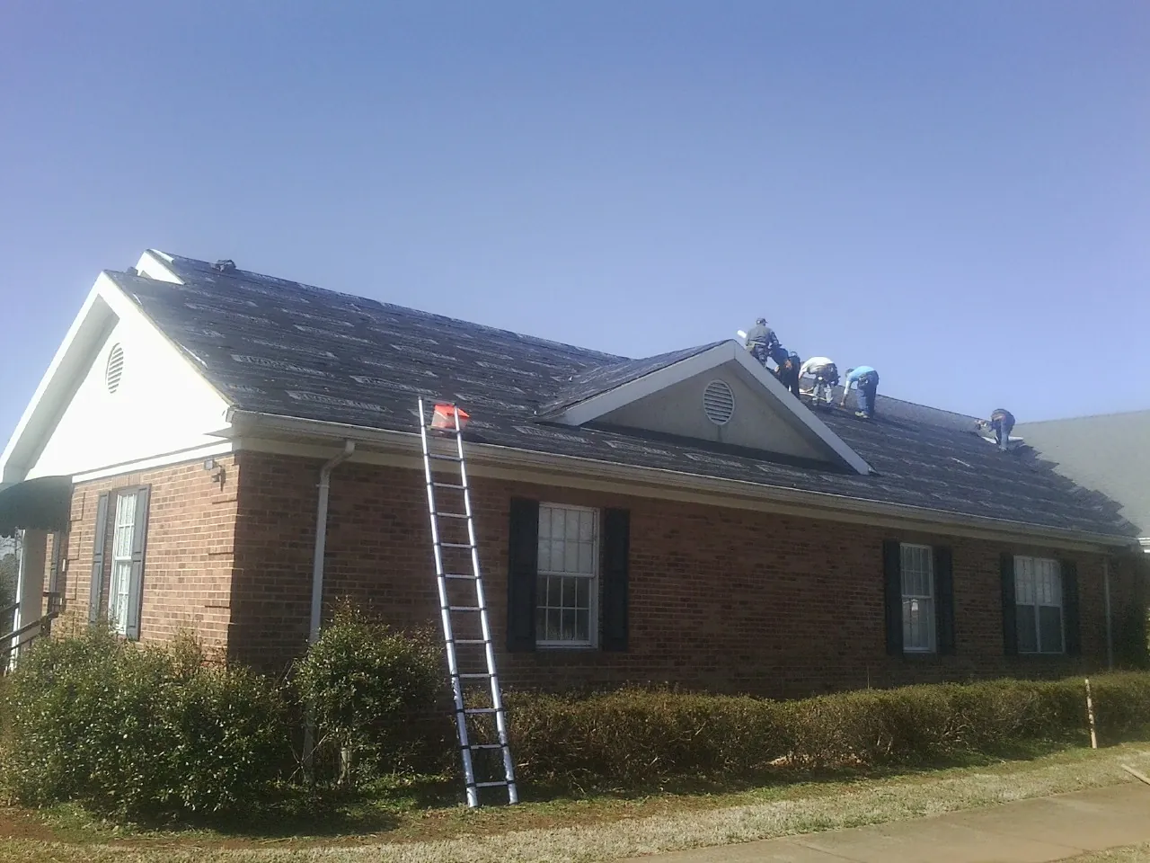 Crew tearing shingles off a brick ranch roof with a ladder set — Emergency Roof Repair work by our crew in Ankeny