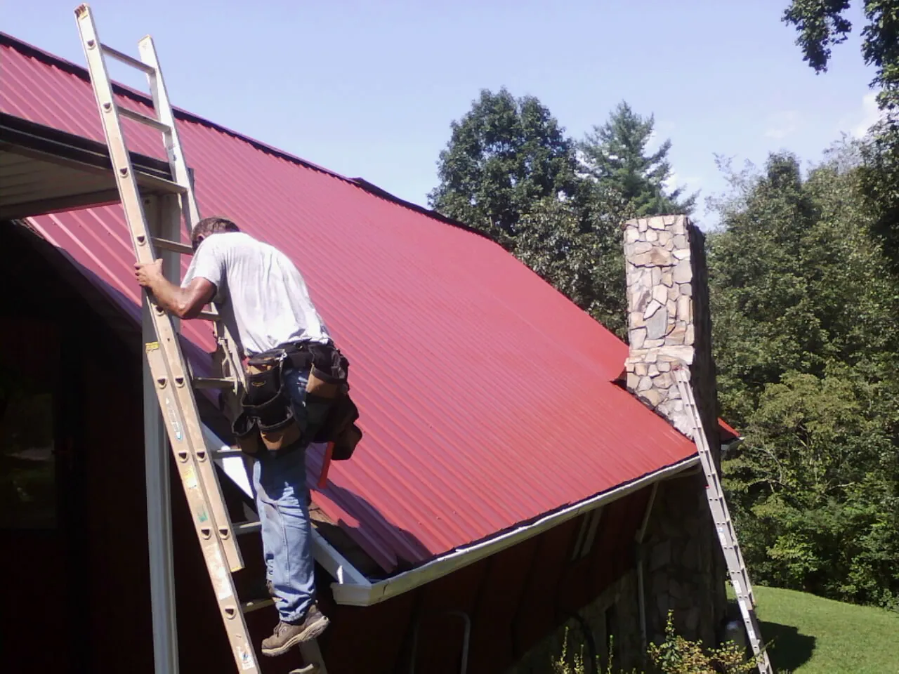 Roofer on a ladder beside a red metal roof and stone chimney — Soffit & Fascia Repair by Roofing Brothers in Ankeny